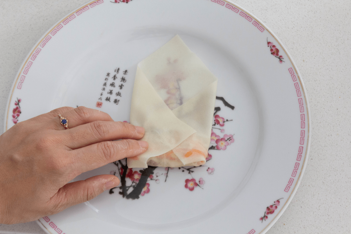 A hand pressing down on a spring roll half wrapped with its left and right corners folded in.