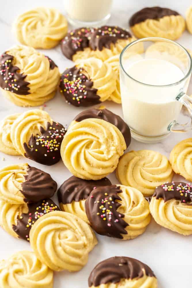 A batch of butter cookies, some chocolate coated, next to a glass of milk