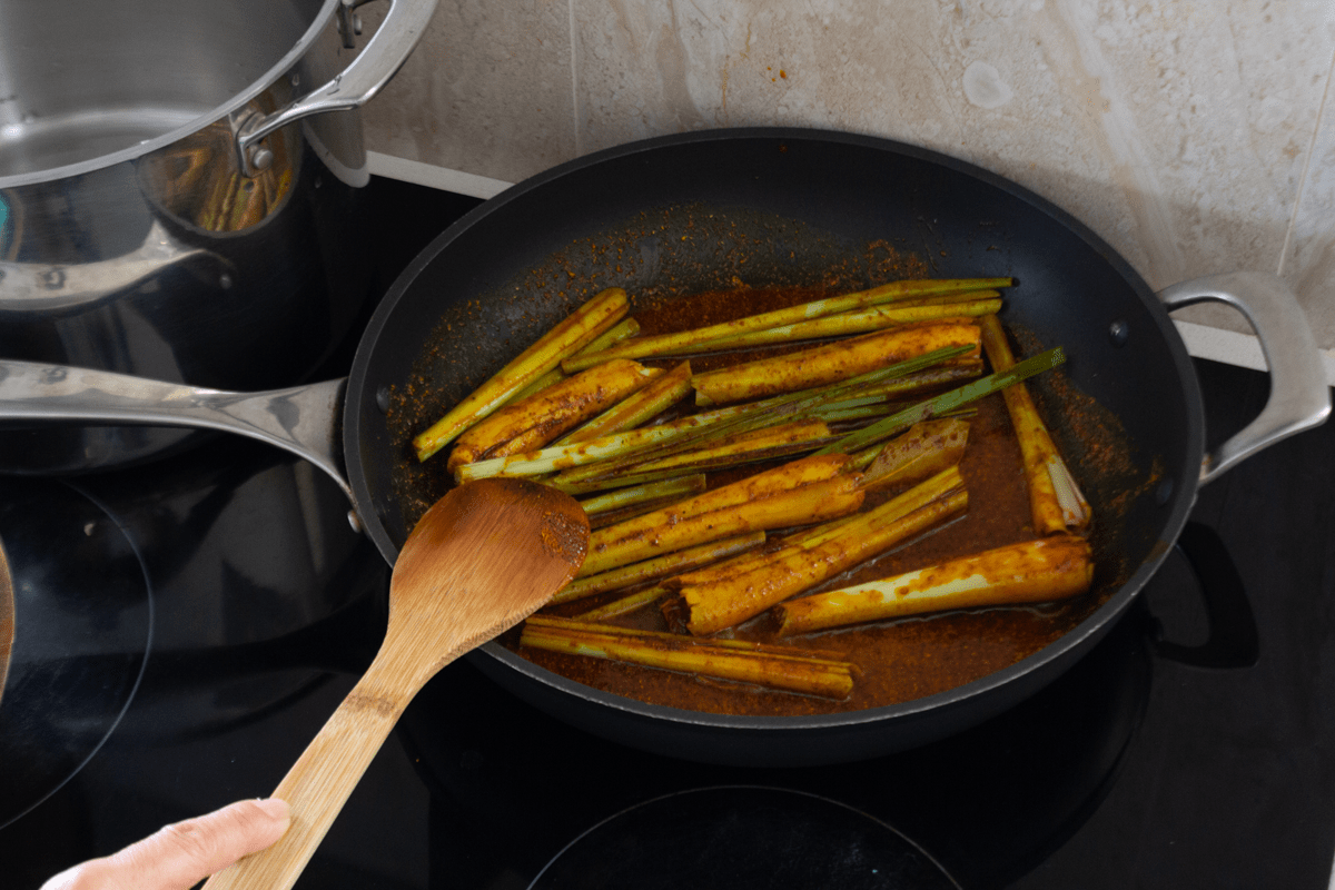 Lemongrass segments in a pan with curry powder and a wooden spoon.