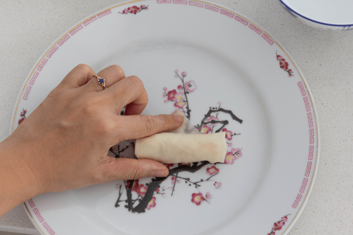 A hand holding an uncooked spring roll with the index finger touching the top corner of the wrapper.